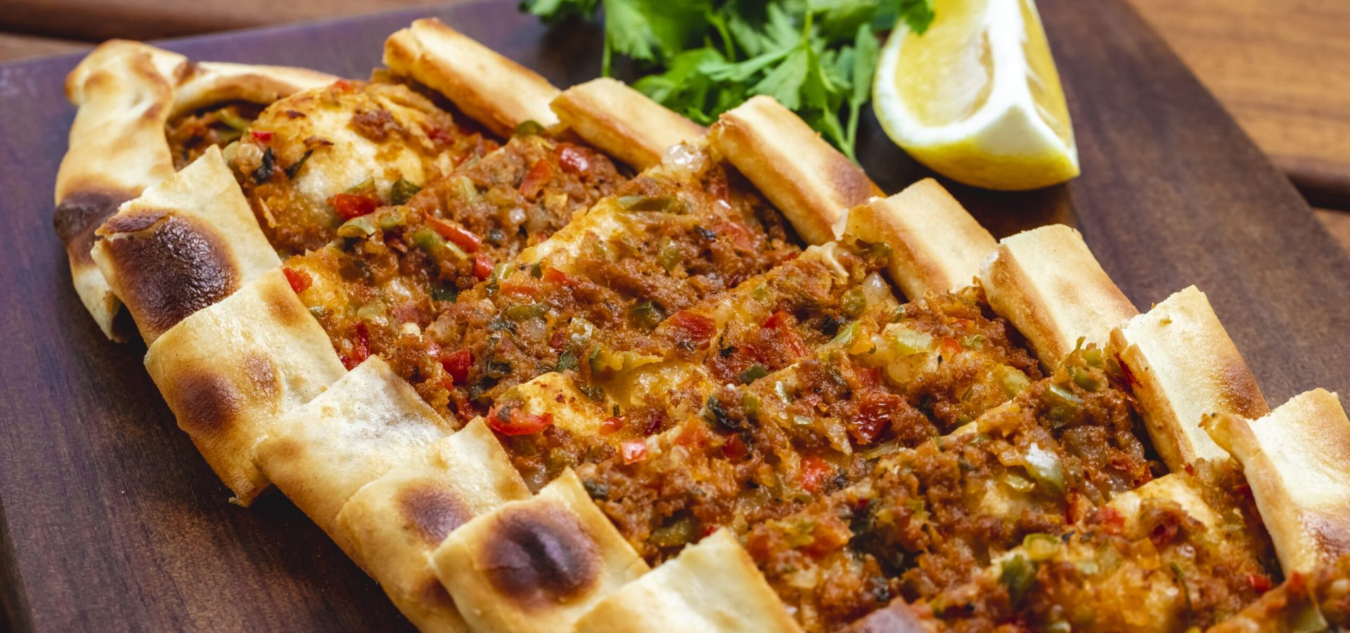side view pide with ground meat tomato onion hot green pepper greens and slice of lemon on a tray